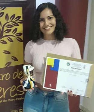 Encontro de Saberes UFOP 2023: estudante do CSI Lab, sorridente, segura o troféu e o certificado de melhor trabalho de iniciação científica diante do banner do evento.